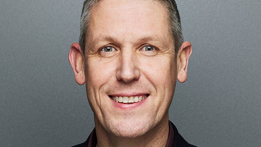 Ein Mann mit kurzen, grauen Haaren lächelt in die Kamera. Er trägt ein weinrotes Hemd und einen schwarzen Blazer.