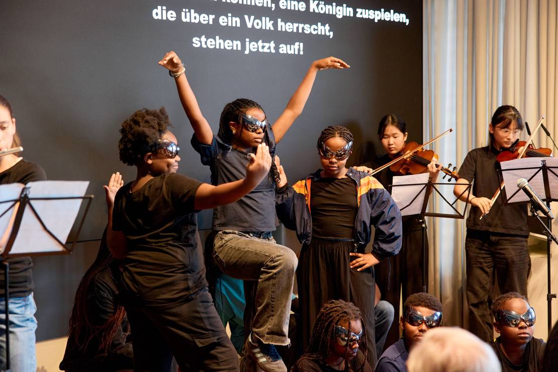 Fotos von Kindern auf der Bühne. Eine hebt die Arme in einer drohenden Geste. Sie tragen große schwarze Sonnenbrillen.