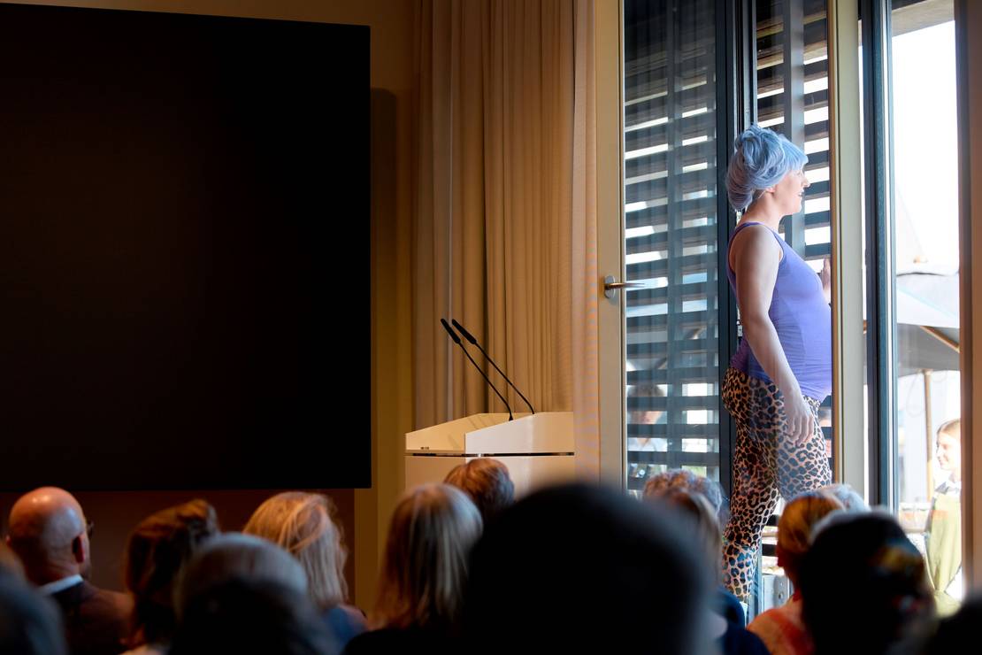 Eine Frau mit lila Perücke singt, während sie aus einem Fenster klettert.