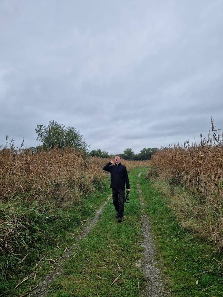 Ein Mann steht auf einem Feldweg zwischen Schilf und hält eine Hand horchend ans Ohr.